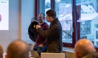 Deux hommes se tiennent devant un public. L'un des deux hommes montre un casque.