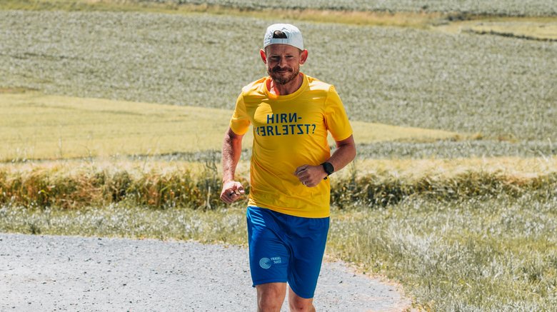 Ein Mann läuft auf einer Landstrasse. Er trägt ein weisses Cap, ein gelbes FRAGILE-T-Shirt und blaue kurze Hosen mit dem FRAGILE-Logo.