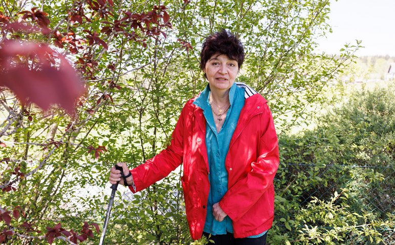 Eine Frau mittleren Alters mit kurzen, dunklen Haaren blickt in die Kamera. Sie trägt eine blaue Bluse und eine rote Jacke. In der rechten Hand hält sie einen Stock. Sie ist von grünen Sträuchern und Pflanzen umgeben.