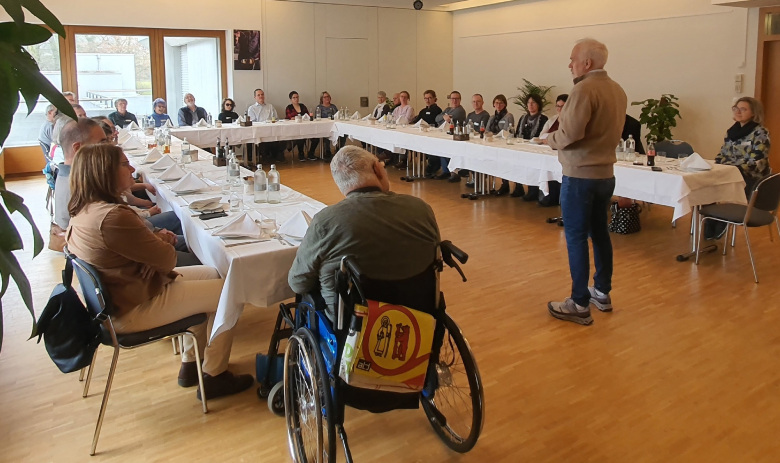 Personen sitzen an gedeckten Tischen in einer U-Form angerichtet. In der Mitte steht eine Person und spricht zu allen.
