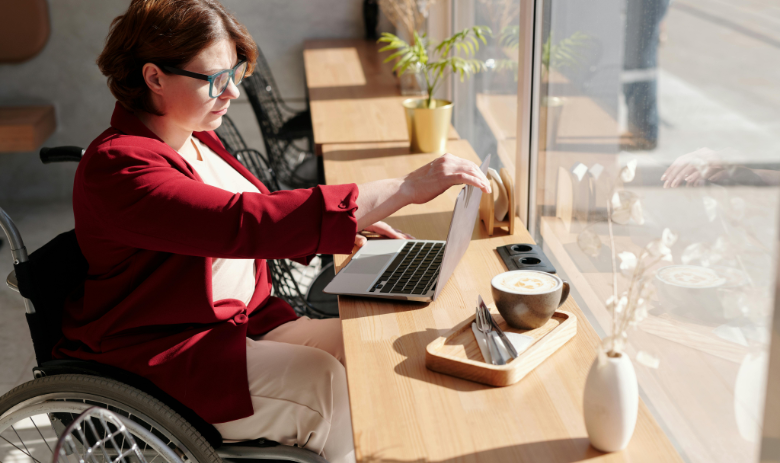 Fachartikel Eine Person im Rollstuhl sitzt an einem Tisch und klappt einen Laptop auf. Daneben steht eine Tasse Kaffee.