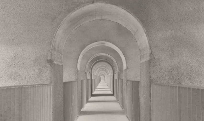 «Gallerie» von Paul Christener Ein schwarz-weiss-Foto eines langen Korridors. Auf beiden Seiten sind Geländer aus Metall, unterbrochen Wandabschnitten mit gewölbten Decken.